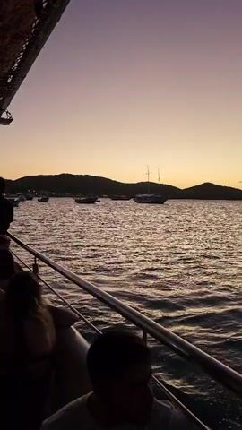 The video begins with a shot from a boat sailing on a body of water at dusk, with landmasses visible in the distance. The camera then quickly pans to the right, capturing another island.  A jet ski zips through the water near the island. The camera continues panning right, showing more distant land, and finally ends with a crowded scene of people on the boat. The group is densely packed, and the lighting indicates it's evening.  Electronic music is playing.