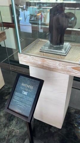 The video shows a dark, irregular piece of wood displayed in a glass case. The wood is mounted on a transparent, multi-tiered pedestal. The case sits on a light-colored wooden stand. In front of the case is a black sign that reads "WITNESS by Caroline Landolfi-Rozea TIME." The sign provides a description of the object, noting it originated from the slopes of Mount Patuha, a stratovolcano in Indonesia, and is a unique type of wood developed through specific circumstances. It also notes that "NATURE IS THE BEST HEALER" and lists "PRICE ON OFFER". The camera moves slightly to show different angles of the object and the sign. Reflections in the glass case show the surrounding environment, including people walking by and other displays.