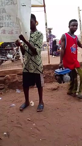 The video shows a street scene. A man in a green patterned shirt and black shorts stands holding a white plastic bag up in front of his head.  A man in a red sports jersey walks past carrying a small blue generator. The camera then moves under a partially covered structure and focuses on a person in a blue shirt working with a black box with wires plugged into it, which is on top of a makeshift wooden table.  In the background, there are motorcycles and other people. The ground is dirt.