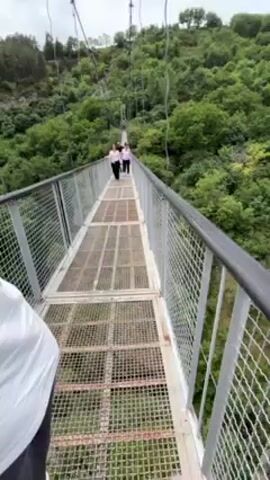 The camera travels along a narrow suspension bridge with gray metal fencing on both sides and a deck made of evenly spaced metal grid panels, offering a view of the gorge below.  Two women are walking towards the other side of the bridge, wearing similar outfits of light-colored tops and dark pants. The camera is pointed forward, capturing the lush green landscape below and on the other side of the bridge, which slopes upward. Beyond the dense trees, there's a craggy cliff face with what appears to be old cave dwellings. The sky above is overcast, with clouds partially obscuring the bright sky.