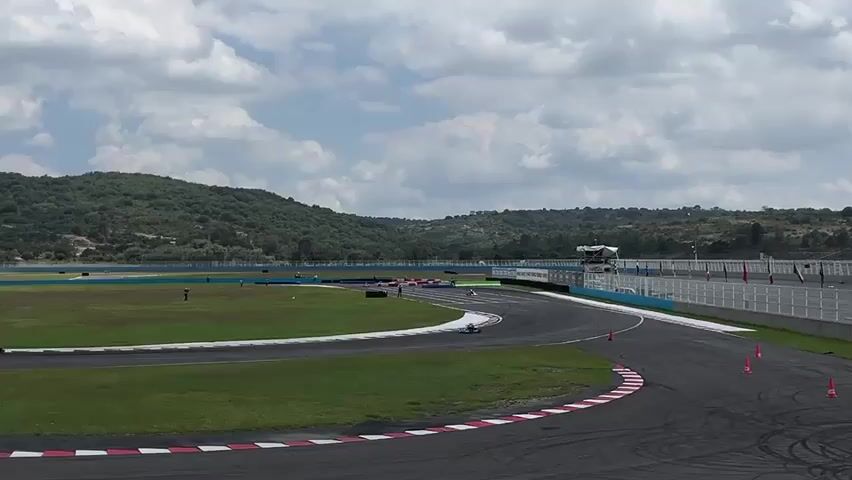 The scene is at a racetrack on a partly cloudy day. The track has black asphalt, marked with red and white curbing. There are several orange cones along the side. There is an area of green grass and a grassy field with some people spread around. A grey wall is on one side of the track. In the distance, a large hill covered in trees is visible. Multiple go-karts are driving on the track. The karts appear to be racing. The camera is stationary.