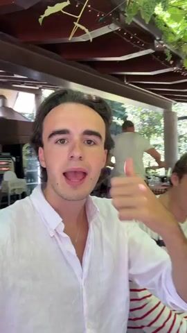 The video opens with a young man in a white linen shirt smiling and giving a thumbs-up towards the camera. He is outdoors at a restaurant with a checkered tablecloth and grapevines overhead. In the background, other people are seated at tables, some laughing and talking. The man gestures to his left, pointing out the location. The scene transitions to show a table laden with plates and glasses, with people engaged in conversation and enjoying a meal. The man continues to speak to the camera, sharing his location. The video ends with a shot of a young woman laughing and a man in a striped shirt covering his eyes.