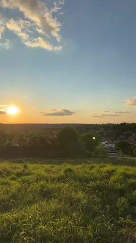 The video shows a slow pan to the right across a landscape. In the foreground, there is a grassy hill bathed in the warm light of a setting sun, which is visible on the far left of the frame. In the mid-ground, there is a small town with red-tiled roofs and scattered trees. A road is visible running through the town. The background consists of rolling hills covered in trees. The sky is a clear blue with a few scattered white clouds, and the sun is casting long shadows. The overall impression is one of peaceful rural life at dusk.