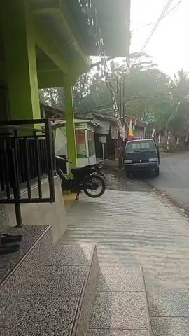 The video shows a street scene in what appears to be a rural or suburban area. On the left side of the frame, there is a building with a green awning and a tiled staircase leading up to it. A motorcycle is parked near the building. The street itself is paved and curves to the right. Several small buildings or shops line the street, adorned with red and white flags. A dark-colored van is parked on the side of the road. Multiple motorcycles are seen on the street, some with riders and some without. One motorcycle in the foreground has two riders. Trees, including palm trees, are visible in the background, along with telephone poles and electrical wires. The sky is overcast. The overall impression is a quiet, everyday scene in a community.