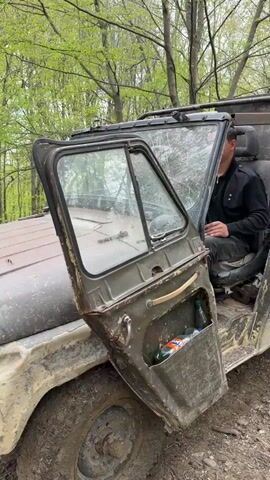 Two men are seated inside a heavily-used, military-green off-road vehicle, likely a UAZ-469, in a forest in Manavi, Georgia. The vehicle is splattered with mud and dirt, suggesting it has been driven through challenging terrain. The driver's side door is open, revealing makeshift storage containing bottles.

The setting is a lush forest with tall trees and vibrant green foliage, indicating late spring or early summer. The ground beneath the vehicle is dirt and mud. The vehicle is stationary, but the sound of its engine can be heard.

The driver, wearing a black top and dark pants, is behind the steering wheel, while the passenger, in a grey shirt, sits beside him. Both men appear calm and focused, looking ahead or slightly to the side. The scene conveys a moment of rest or preparation before continuing an off-road journey through the natural landscape. No visible text is present.
