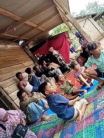 A group of people are gathered indoors, possibly in a damaged structure. They are sitting on a colorful mat laid out on a wooden floor. Some are leaning against the wooden walls. The roof appears to be made of corrugated metal, with some exposed beams. A red fabric is hanging in the background. The people, including men, women, and children, are looking in the same direction, suggesting they are watching something. The overall scene conveys a sense of shared attention and possibly concern or anticipation.