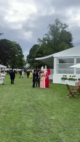 The video shows a group of people dressed in formal attire, including top hats and elaborate dresses, standing on a grassy lawn. In the foreground, two men in black top hats and tailcoats are facing a woman wearing a large, pink ruffled dress and a feathered headdress. Another woman in a red dress and a red headdress stands next to the woman in pink. They appear to be engaged in conversation. In the background, more people are walking around, and there are tents and trees visible. The overall scene suggests an outdoor event, possibly a race meeting or a formal garden party.