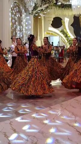A group of women in ornate, brightly patterned dresses with full skirts are dancing in a circle on a polished marble floor. A man in a white shirt and dark pants is standing in the center of the circle, facing the camera and clapping. The room is elaborately decorated with draped fabrics, floral arrangements, and chandeliers. Other people are visible in the background, seated at tables or standing. The lighting is dynamic, with colored spotlights projected onto the floor, creating geometric patterns. The women are performing synchronized movements, with their arms extended and their skirts flowing.