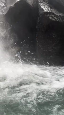 The video shows a close-up view of a waterfall cascading over dark, jagged rocks. The water is white and turbulent, creating a foamy pool at the base of the falls. The camera angle is low, emphasizing the power and movement of the water as it crashes down. The surrounding environment appears to be a rocky, natural setting.