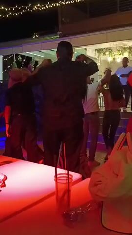 A group of people are dancing at night on an outdoor patio decorated with string lights. In the foreground, a person in a black shirt and pants is raising their hands, seemingly dancing. To the right, a person in a red shirt is dancing with a woman in a black top and patterned pants. In the background, several other individuals are visible dancing and socializing on a tiered platform. Tables with drinks are set up in the foreground. The overall atmosphere appears to be a lively social gathering.