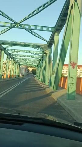 The video shows a car driving across a bridge. The bridge has a green metal truss structure and an orange railing. On the other side of the railing is a body of water. In the distance, there are buildings and trees. The sky is blue and clear. There are other cars on the bridge and some people walking on the sidewalk. The video is filmed from inside the car, looking out the windshield. The sun is shining and casting shadows on the bridge.