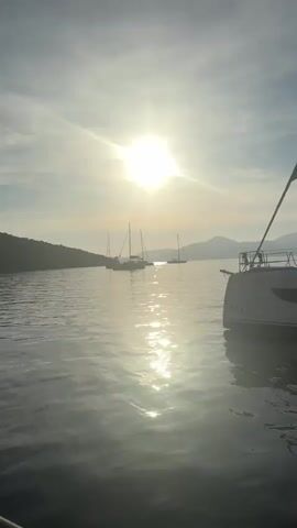 The video captures a serene waterscape, likely a bay or inlet in Bigova, Montenegro, at either sunrise or sunset. The sun is a prominent, bright orb in the sky, casting a strong, sparkling reflection across the calm water. Several sailboats and catamarans are visible in the middle distance, appearing as silhouettes against the bright sky and distant, hazy hills or mountains. In the foreground, the bow of a catamaran is partially visible on the right side of the frame, with the word "BALI" clearly discernible on its hull. The camera appears to be on a gently moving boat, providing a low-angle perspective of the tranquil scene. The sky is bright with some light cloud cover, contributing to the soft, atmospheric lighting. Ambient human speech can be heard in the background.