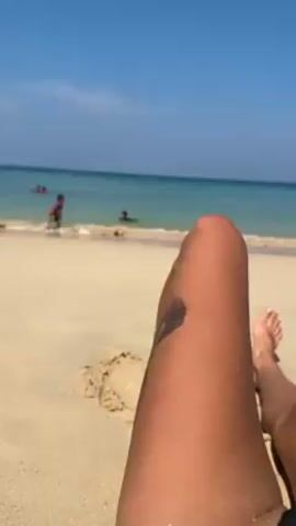 The scene is a sunny beach with a clear blue sky and turquoise water. The camera focuses on the lower legs of a person lying on the sand. The person has tanned skin and a small tattoo on one leg. In the background, several people are in the water or walking along the shoreline. There is a small sandcastle to the side. The scene is calm and relaxing.