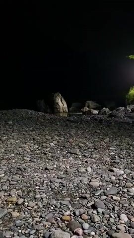 The video shows a rocky shoreline at night. A light source illuminates a portion of the scene, casting shadows and highlighting the texture of the stones. In the background, large rocks are visible, and a body of water appears to be present. A rudimentary structure, possibly a table or bench, is visible in the distance. The overall atmosphere is dark and quiet.