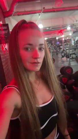 A young woman with long brown hair is standing in a gym, the lighting is red. She is wearing a black and white sports bra and black shorts. In the background, there are dumbbells and gym equipment. She is looking at the camera and smiling.