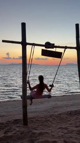 A person sits on a swing on a beach. The swing is made of wood and suspended from a bamboo frame. The person is facing away from the camera, towards the ocean. The sun is setting, casting a warm glow over the water and the sky. The person is wearing a light-colored outfit. The waves gently lap at the shore. The sand is light in color. The sky is painted with hues of orange, pink, and purple. Islands or landmasses are visible on the horizon. A sign is hanging from the bamboo frame above the swing. The person's hair is long and appears to be tied back. The person's feet are visible, dangling just above the sand. The person is holding onto the ropes of the swing. The overall scene evokes a sense of peace and tranquility.