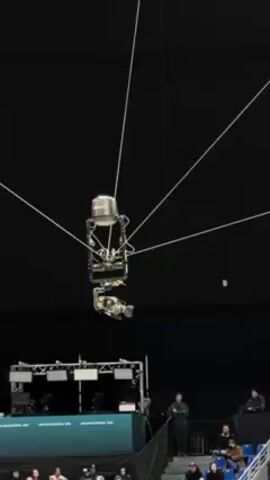 The video shows a robotic camera suspended in the air over a tennis court or stadium. It is held up by a system of wires attached to the ceiling. Below the camera, there are spectators watching the game. In the background, there is a large screen that reads "MILANO CORTINA 2026." The camera appears to be tracking the action on the court.
