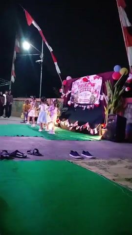 This is a night-time video of a group of children performing on a stage outdoors. The stage is decorated with red and white banners and balloons. Three young girls in dresses are on the stage, appearing to dance or perform. There is a speaker on the right side of the stage. In the foreground, there is a green mat and some pairs of shoes. Two adult figures are visible in the background on the left. The scene appears festive, possibly for a local event or celebration.