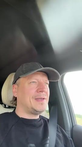 A man wearing a black baseball cap and a black t-shirt is sitting in the driver's seat of a car. He is looking at the camera and speaking. The interior of the car is visible, including the headrest of the passenger seat and the ceiling of the car. Outside the car, a field of grass is visible. The man smiles and touches his cap.
