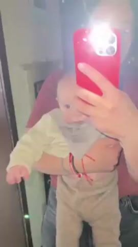 A person wearing a dark red shirt and gray jeans is holding a baby in their arms. The baby is wearing a beige sweater, a gray bib, and brown corduroy pants. The person is using a red smartphone to take a picture of them and the baby in a mirror. The flash on the phone is illuminated. The baby's stomach is exposed and slightly red. A doorway or entrance is visible in the background.