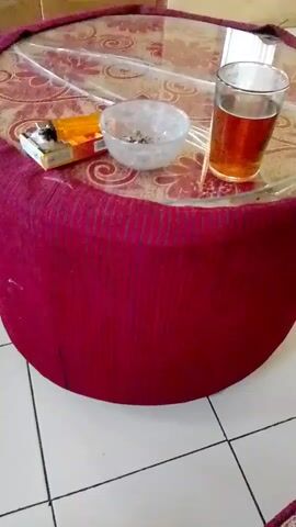 The video shows a close-up of a round coffee table covered with a patterned cloth and a clear plastic cover. On the table, there is a pack of cigarettes, an ashtray with cigarette butts, and a glass of amber liquid, likely tea. The table is situated on a tiled floor next to a sofa with a similar patterned fabric. The background includes a window with white curtains and a light-colored wall adorned with two framed pictures and three small white shelves holding potted plants. The camera pans slightly to the right, revealing more of the sofa and wall.