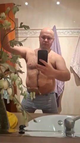 A shirtless, bald man in blue boxer briefs takes a selfie in a bathroom mirror. He stands in front of a sink and countertop with a vase of flowers on the left.  Behind him are tiled walls with a couple of towels hanging on a towel rack.  He poses with his right arm raised in a flexing motion. The video is shaky due to him holding the phone.