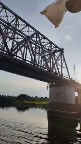 The video captures a low-angle shot of a metal truss railway bridge spanning a body of water. The perspective is from a boat moving along the water, as indicated by the slight ripple effect on the surface. Sunlight is visible in the background, creating a glare and casting long shadows. The bridge is constructed with a complex network of beams and supports. On the far side of the bridge, a grassy bank with scattered trees is visible. In the upper right corner of the frame, a white object, possibly part of a boat or other structure, is partially visible. Small dark specks, likely birds, can be seen in the sky. The video is a short, still clip with no discernible action or interaction.