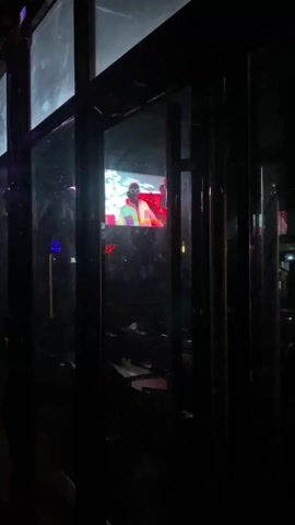 The video shows a dark interior with a window-like structure dividing the view. Through the window, a screen displays video of a man wearing a pink hat, white shirt, and white-framed sunglasses. He is smiling and appears to be singing or rapping. Other people are faintly visible behind him in the video. The overall setting seems to be a bar or club with low lighting.