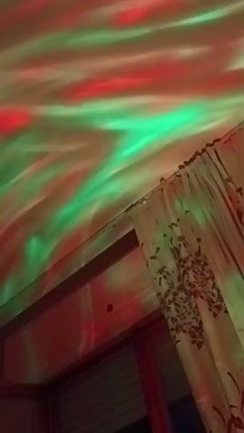 This short video showcases a ceiling illuminated by a light projector. The projector displays swirling patterns of various colors, resembling an aurora borealis. The colors cycle through green and red, then purple and blue, followed by a bright blue, and finally a mix of purple and pink. The scene appears to be indoors, with a window and curtains visible on the right side of the frame. The lighting suggests it could be nighttime or a dimly lit room, enhancing the visual effect of the light display. There is no visible text within the video. The overall mood is tranquil and mesmerizing, created by the dynamic and colorful light patterns.