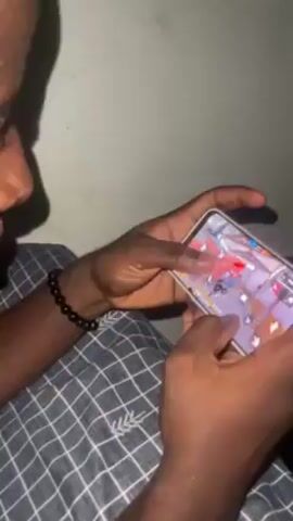 Here is a description of the video:

A person is playing a mobile video game on a smartphone while lying in bed. The person's hands are visible holding the phone and using their fingers to control the on-screen game elements. The game appears to be a first-person shooter, with the character running through an indoor environment. The person is wearing a bracelet on their left wrist and is lying on a bed with a gray and white checkered pattern. The background is a plain, light gray wall.