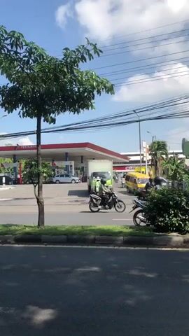 The video shows an outdoor scene with a gas station in the background. A white car and a truck are visible at the gas station. In the foreground, a tree stands tall, and a street runs across the bottom of the frame. On the street, two people are riding a motorcycle, and another motorcycle is visible further down the road. The sky is blue with some clouds, and power lines are visible overhead. The overall impression is a busy street scene on a clear day.