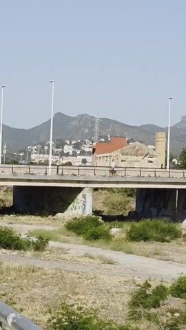 The video presents a series of daytime scenes from an urban or suburban environment under a clear, sunny sky, with mountains visible in the background.

The initial clips (0:00-0:02) show a multi-lane bridge spanning a dry, undeveloped area. Cars are seen driving across the bridge, and a person rides a bicycle from right to left. In the background, green-brown mountains are dotted with buildings and what appears to be an industrial structure or communications tower.

The subsequent clips (0:02-0:05) transition to street-level views. A paved sidewalk runs alongside a street where several cars are parked. A person is visible walking on the sidewalk. The street features a curve leading to a roundabout, adorned with trees, including prominent palm trees. Other cars are seen driving on the road and around the roundabout. Buildings line the streets.

Notable details include the bright, clear daylight conditions throughout the video. A blue pedestrian crossing sign with a white figure is visible in one frame (0:04-0:05), partially obscured by a green recycling bin. The license plate "5255 GCD" is clearly visible on the rear of an orange hatchback car in multiple shots (0:03-0:05). Some text, likely instructions or categories for recycling, is faintly visible on the green bin but is not fully legible. The overall setting suggests a warm, dry climate.