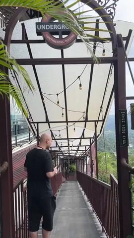 A bald man wearing a black t-shirt and black shorts stands on a covered outdoor walkway with metal railings and a transparent roof adorned with string lights. Above him, a sign reads "UNDERGROUND" with "CHARCOAL GRILL" and "CONTINENTAL CUISINE." Another warning sign is visible on a support beam to the right. The walkway offers a view of distant green mountains and a valley. The man holds a smartphone, appears to take a picture, briefly smiles at the camera, then lowers his phone to look at it. Palm fronds are visible on the left and right sides of the frame.