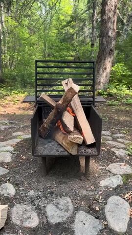 This media file showcases a close-up view of a campfire in a wooded setting, likely a campground. The primary object is a metal fire pit containing burning logs arranged in a teepee-like structure. Flames are visible, licking at the dry wood, and a wisp of smoke drifts upwards. The fire pit itself is set on legs and has a grilling grate attached to the back. The ground surrounding the fire pit is covered in gravel and scattered with large, natural stones. In the background, lush green trees suggest a temperate climate and daylight hours, possibly late spring or summer. The overall atmosphere is serene and rustic, evoking a sense of outdoor recreation and relaxation. There is no visible text in the media.