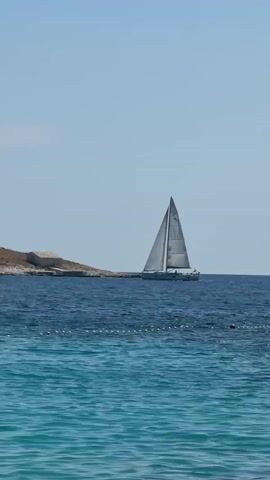 A sailboat with white sails is visible on a deep blue body of water. To the left of the sailboat, a small, rocky island with a white structure on it is present. The water in the foreground is a lighter, turquoise color, with small waves. The sky is clear and pale blue. The scene appears to be a sunny day.