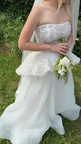 A young woman in a white strapless wedding dress with a beaded bodice and a peplum is holding a bouquet of white flowers and greenery. She is wearing a veil and earrings. The woman is standing outdoors in front of a green hedge. The video shows the woman from the waist up, as she smiles and looks down at the bouquet.