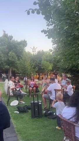 A group of people are gathered in a grassy outdoor area, seated in chairs in a circle. They are playing hand drums. Some people are holding the drums on their laps, while others have them placed on the ground. There are trees and foliage in the background, suggesting a park or garden setting. The sky is visible above the trees and appears to be clear and bright. In the distance, a person is visible standing on top of a structure, their arms raised. A portable speaker is situated in the center of the circle. Several bags and a hat are on the grass around the chairs. The overall atmosphere appears to be a casual gathering with music.
