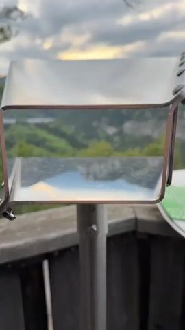 The video shows a landscape view of green mountains and a cloudy sky, reflected in a metal mirror-like viewing device. The viewing device is mounted on a metal pole attached to a wooden railing. The reflection in the mirror is clear and shows the details of the landscape, including the mountains, trees, clouds, and a touch of sunset.