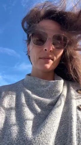 A woman with long brown hair and sunglasses is shown from the chest up against a bright blue sky. She is wearing a gray sweater with a textured knit. Her hair is blowing in the wind, partially obscuring her face. The camera is angled upwards towards her.