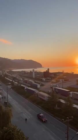 The video shows a sunset over a coastal area. In the foreground, there is a road with a few vehicles, including a red car and a person walking. Further back, there is a row of parked semi-trailer trucks. Beyond the trucks, there is a body of water, likely the sea, and hills in the distance. The sun is low on the horizon, casting a warm orange glow over the scene. There are some industrial structures and buildings visible near the water. The sky is a gradient of blue and orange.