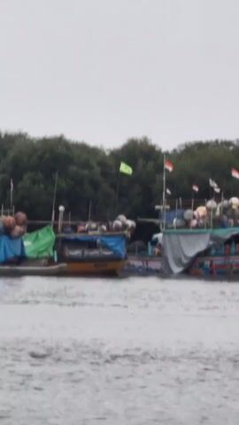 A still shot shows two fishing boats docked side by side on a body of water. The boats are filled with round, tan-colored objects, possibly buoys or fishing floats. Both boats have flags on poles. One boat has a green and yellow flag, while the other has several Indonesian flags. The background is a dense line of green trees, suggesting a coastal or riverine environment. The sky is overcast.