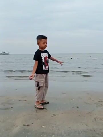 A young boy stands on a sandy beach in Kualaidi, Indonesia, under an overcast sky. He is wearing a black T-shirt with a "SPIDER-" logo, light brown cargo pants, and open-toed sandals.

At the beginning of the video, the boy faces slightly left, extending his right hand forward with an open palm. He then bends at the waist, looking down at the sand. He briefly straightens before fully crouching, bringing his left hand to rest on his knee as he uses his right hand to pick something up from the sand.

The calm sea forms the background, with gentle waves rolling onto the shore. A small boat is visible in the distance on the left side of the frame. The sand is light brown and appears slightly damp near the waterline. The overall impression is one of quiet exploration on a cloudy day by the sea.