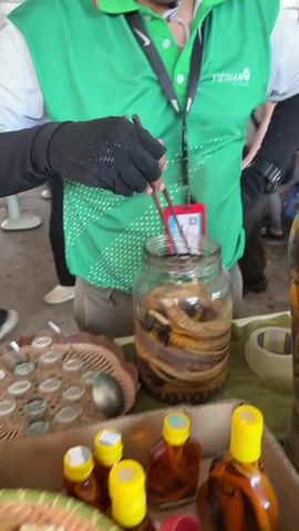 In a market or stall in Thới Sơn, Vietnam, a person wearing a green polo shirt with white polka dots and black sleeves/gloves uses wooden chopsticks to stir the contents of a large glass jar. The jar contains several snakes preserved in an amber-colored liquid, likely snake wine. A red ID card with a QR code and Vietnamese text, including "NGUYỄN NHÀN PHÁT" and "141 160 907", hangs around the person's neck. On the table, to the left, is a tray with several small, empty glasses and a spoon. To the right, smaller bottles with yellow caps, filled with a similar amber liquid, are arranged. Another large jar containing preserved snakes is partially visible on the right. The person's shirt also has "VIETNAM" printed on it. The scene suggests a demonstration or preparation of the traditional beverage.