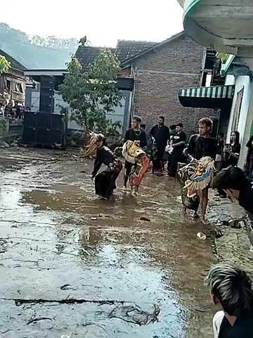 The video shows a performance taking place on a wet, muddy surface in a village-like setting. Several individuals are present, some dressed in black, appearing to be part of the performance or support crew. One performer is dressed in costume and seen dancing in the mud. Other individuals are watching from behind a stone wall. A sound system is visible in the background, likely providing music for the performance. The environment appears to be a village with houses and trees in the background. There is a banner that reads "RYANDA" on the wall.