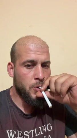 A close-up shot features a man with a bald head and a full, dark beard, looking slightly downwards and to the left. He is wearing a dark-colored t-shirt with the word "WRESTLING" partially visible in white text on the front. He holds an unlit cigarette in his mouth. His right hand holds a blue and black lighter, which he uses to ignite the cigarette. He then takes a drag from the lit cigarette. The background is a plain, light-colored wall. The man's expression is neutral and focused on the act of lighting his cigarette.