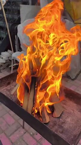 A bonfire is burning in a metal grill. The fire is bright orange and yellow, with flames reaching high into the air. Several pieces of wood are visible within the fire, arranged in a teepee shape. The grill itself is dark metal and sits on a stand. The ground beneath the grill appears to be paved with pinkish-red bricks. In the background, there are some objects covered with white and patterned fabric, as well as a stack of bricks and a metal railing.