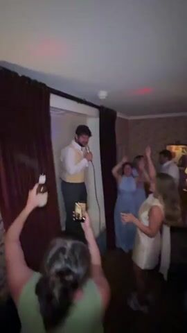 A group of people are celebrating in a room with dim lighting and colored disco lights projected onto the ceiling. In the foreground, a person in a green top is recording the scene with a smartphone. To their left, a man in a white shirt and yellow vest stands on what appears to be a windowsill, holding a microphone and singing. Several people are dancing in the background, including a woman in a light blue dress and a woman in a white dress. A DJ setup with turntables is visible in the lower right of the frame. The room has patterned wallpaper and decorative lamps.