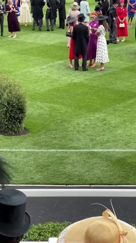 The video shows a grassy field with a white line marking a boundary. Several people are present in formal attire, including hats and dresses. A horse is being led across the field by a handler. In the foreground, two people are visible from behind, wearing hats. One is wearing a black top hat and the other a wide-brimmed straw hat. The overall scene appears to be an outdoor event, possibly a racecourse.