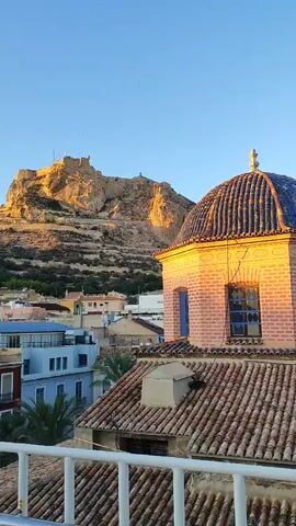 This video shows a cityscape at sunset. In the foreground, there is a rooftop with terracotta tiles and a white railing. To the right, a portion of a building with a dome is visible. The building is made of brick and has arched windows. In the background, there are several colorful buildings and a large mountain with a castle on top. The sky is clear and blue, with the sun casting a warm glow on the landscape. The overall scene is tranquil and picturesque.