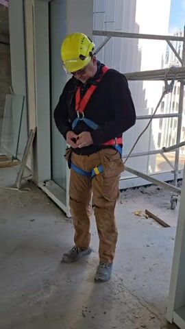 A construction worker in a hard hat, safety glasses, black shirt, and tan pants is clipping the harness around his waist. He is standing in an indoor construction site with glass panels on one side and scaffolding in the background.
